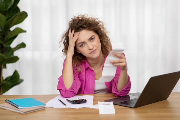 A young adult looking stressed while holding bills and a credit card, with a laptop displaying debt consolidation options in the background.