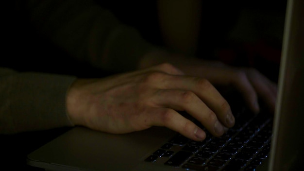 A close-up of hands typing on a keyboard at night, with a shadowy figure in the background, representing the anonymity and hidden nature of online identity theft.